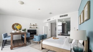 Una habitación de hotel moderna con una cama de madera, un escritorio con sillas, un televisor y un baño visible a través de una puerta de cristal.