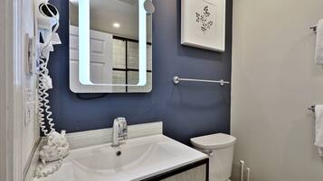 Une salle de bain avec un lavabo blanc, un miroir et des toilettes. On y trouve un porte-serviettes et un tableau encadré accroché au mur.