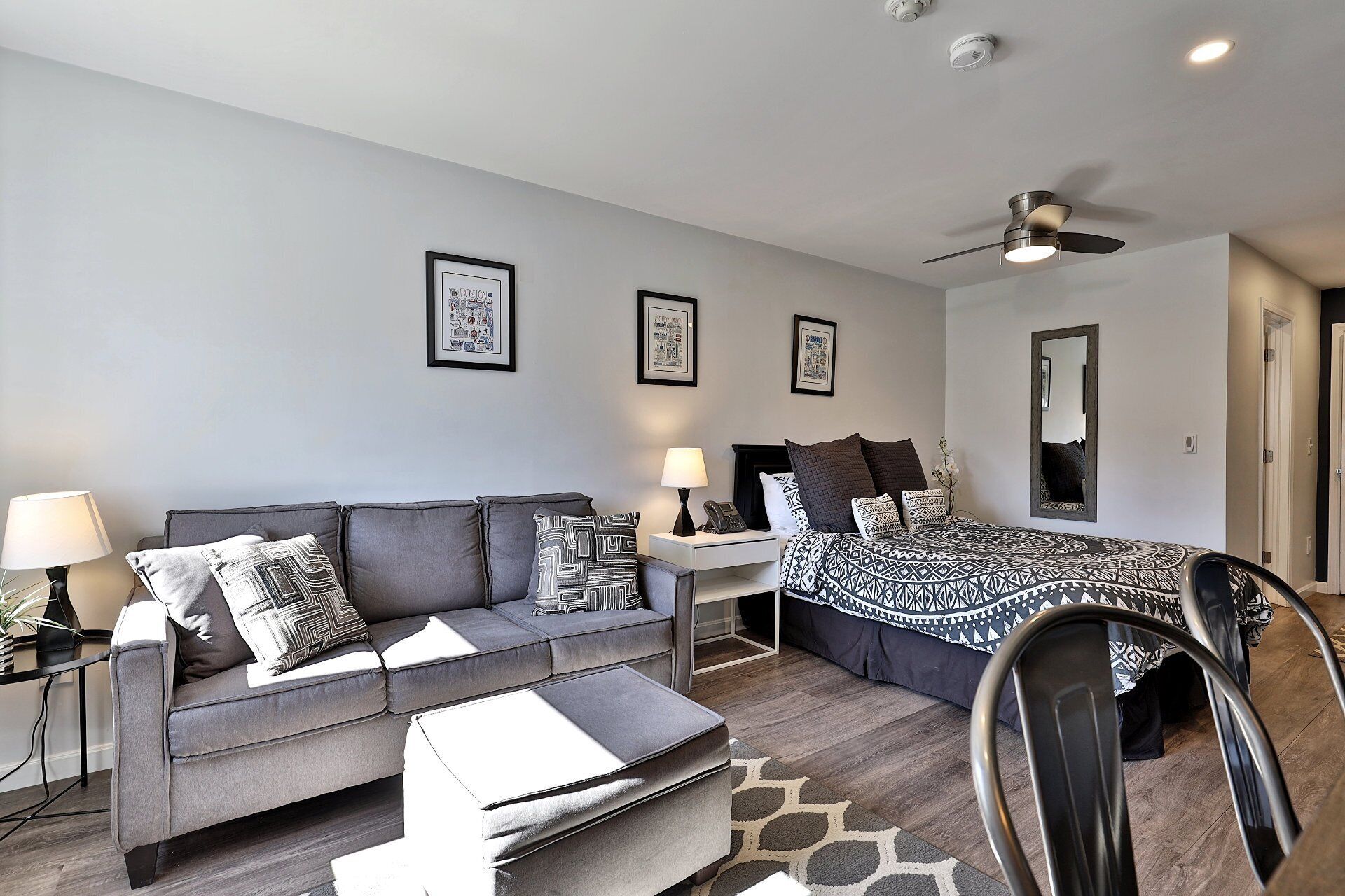 Une chambre moderne avec un canapé gris, un lit avec une literie à motifs, une table de chevet avec une lampe et des tableaux encadrés accrochés au mur.