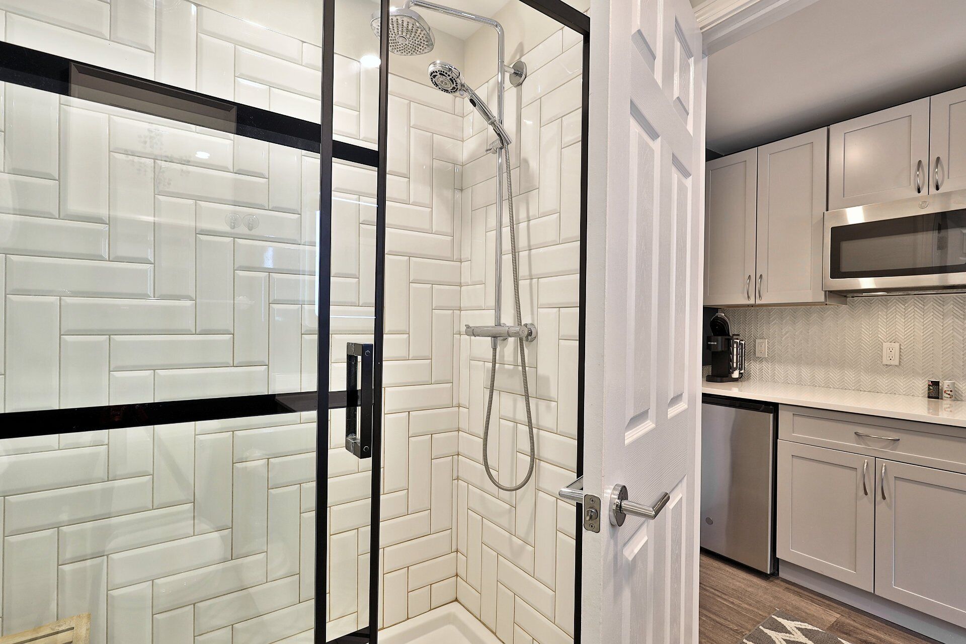 A modern bathroom with a glass shower enclosure, white subway tile walls, and a kitchen area with light-colored cabinets and a microwave.