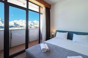 Habitación de hotel con una cama grande, una mesita de noche, una lámpara y vistas a montañas nevadas a través de una puerta corrediza.