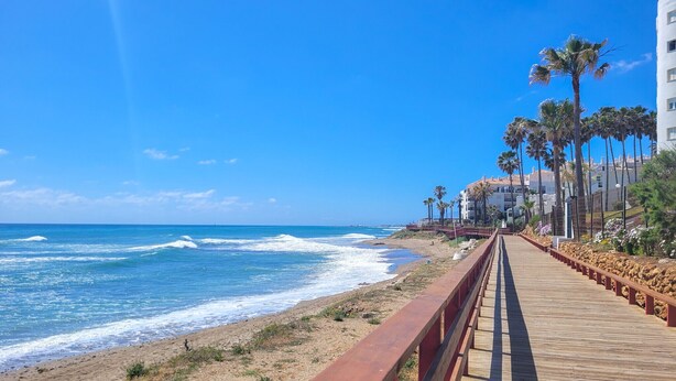 A wooden boardwalk along a beach with ocean views, palm trees, and buildings in the background.