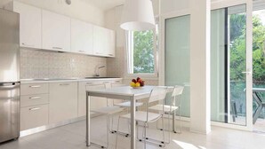 A modern kitchen with white cabinets, a stainless steel refrigerator, a dining table with chairs, and a large window providing natural light. - Holiday apartment with air conditioning and balcony (Lignano Sabbiadoro)
