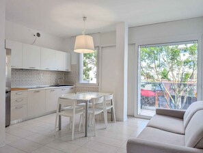 A modern kitchen with white cabinets and a dining area with a white table and chairs. - Holiday apartment with air conditioning and balcony (Lignano Sabbiadoro)