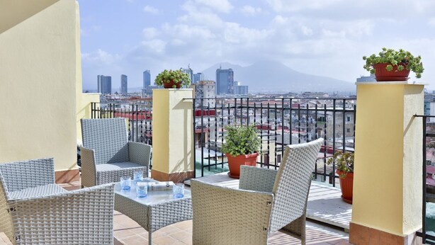 A balcony with wicker furniture, potted plants, and a cityscape view. - GuestHost - This bright, recently renovated apartment is located in a charming 18th-century building, in a quiet and secluded location despite being in a lively and central area just steps from Via Foria and Via Cesare Rosaroll. The Cavour Metro Line 2,  (Naples)