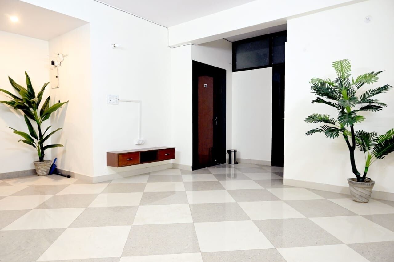 A spacious hallway with a checkered floor, a white wall, and a wooden door. There are two potted plants on either side of the hallway.