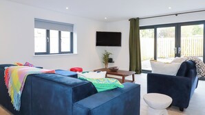 A modern living room with a blue sofa, a white armchair, and a small table. There is a TV mounted on the wall and a window with curtains. - Sea Barn (St Austell)