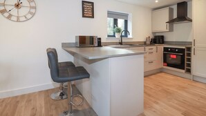 A modern kitchen with a central island, bar stools, a clock, and a sign listing Italian dishes. - Sea Barn (St Austell)