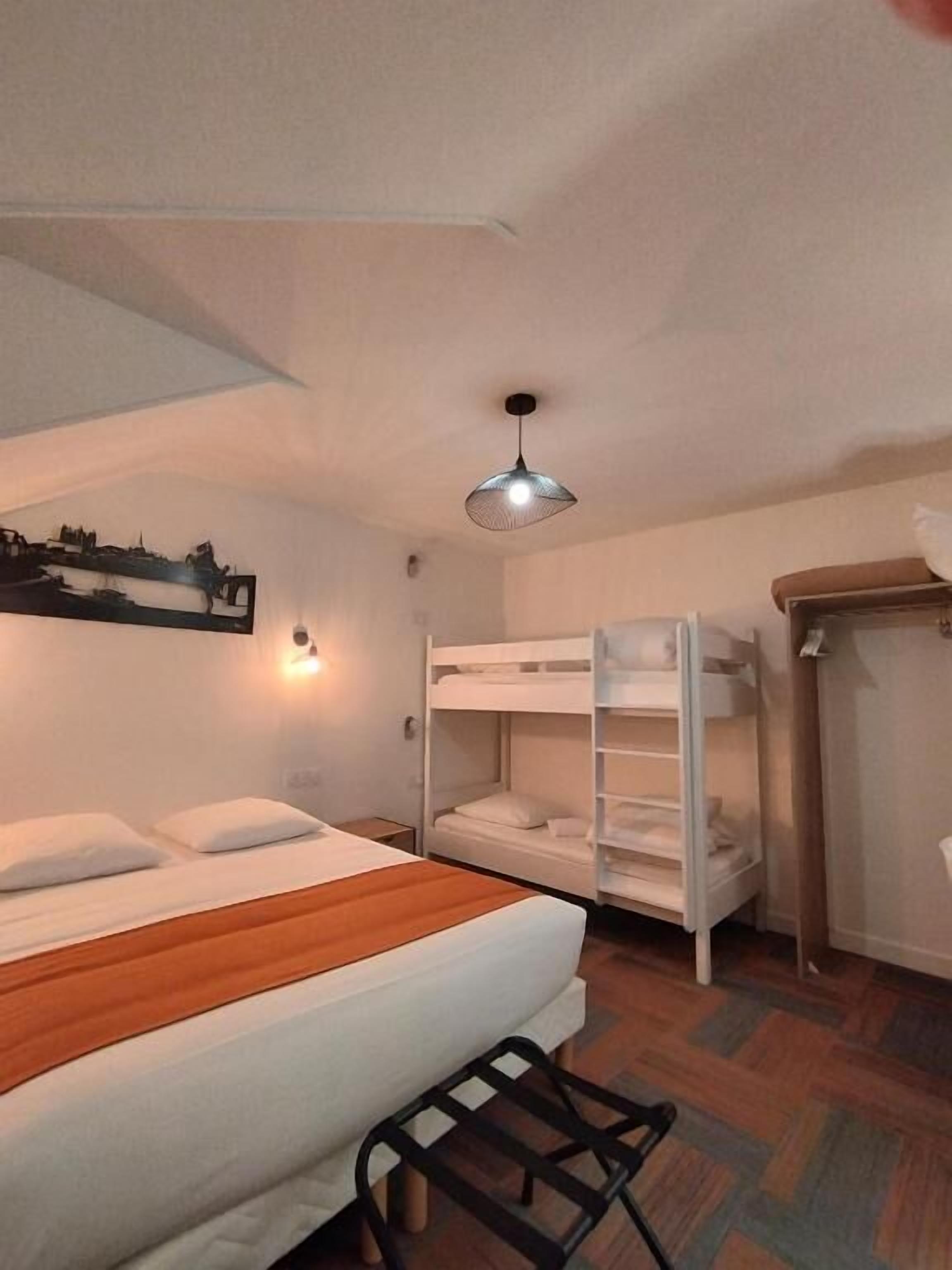 A hotel room with a single bed, a bunk bed, a wooden bench, a wall-mounted light fixture, and a hanging light.