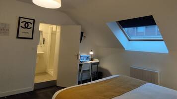 A bedroom with a bed, a desk with a computer, a window, and a door leading to a bathroom.