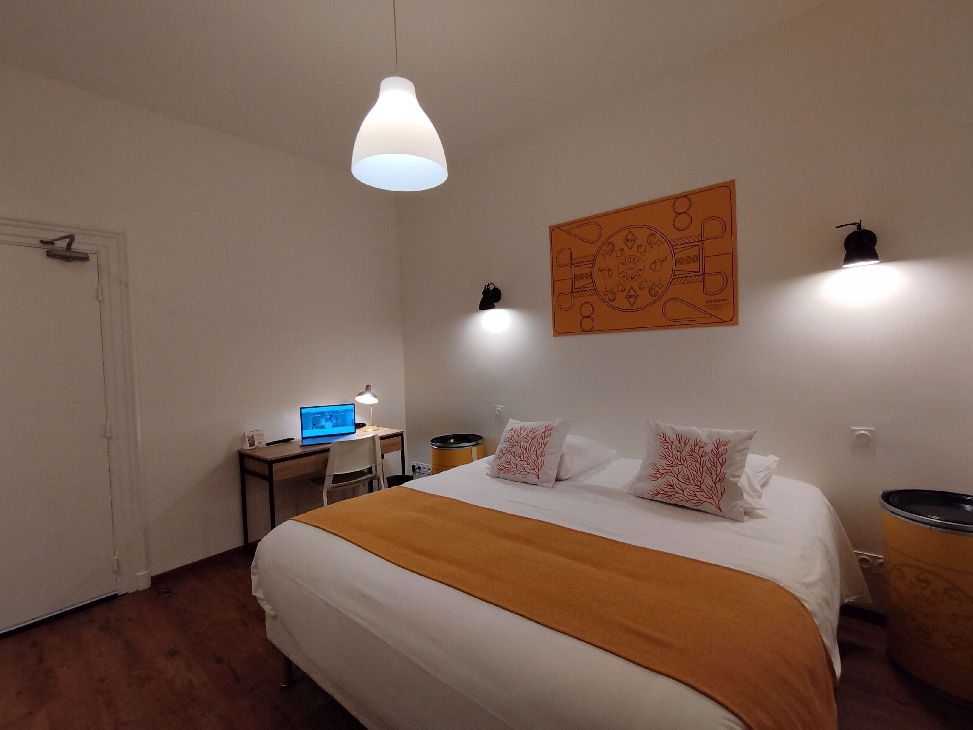 A bedroom with a bed, a desk with a laptop, a hanging light, and a wall art piece.
