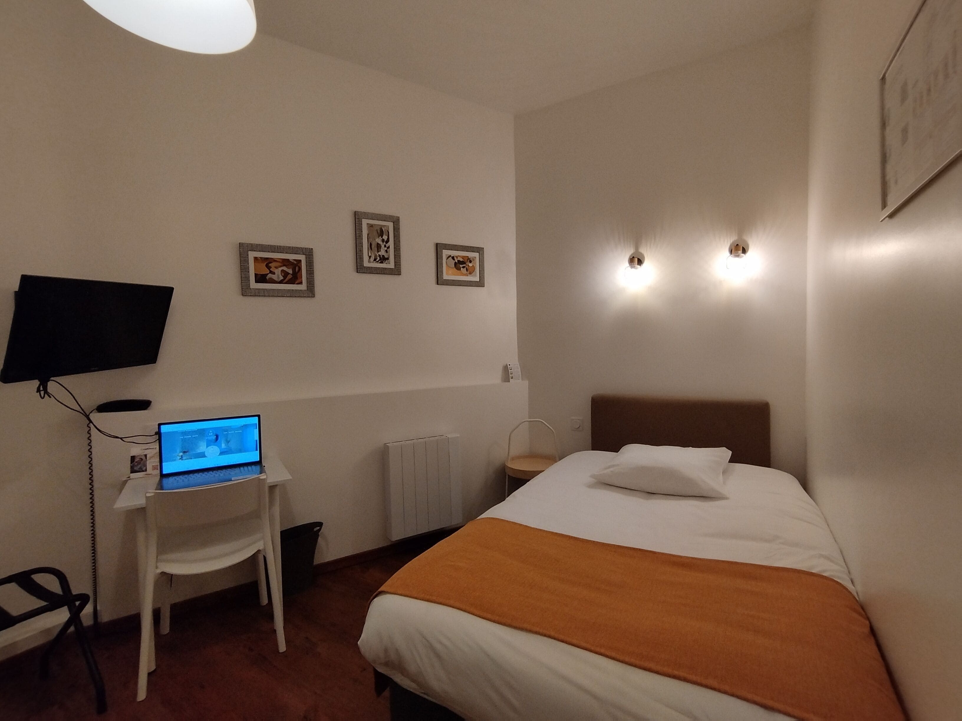 Habitación de hotel con cama, escritorio con una computadora portátil, silla, televisor y tres cuadros en la pared.