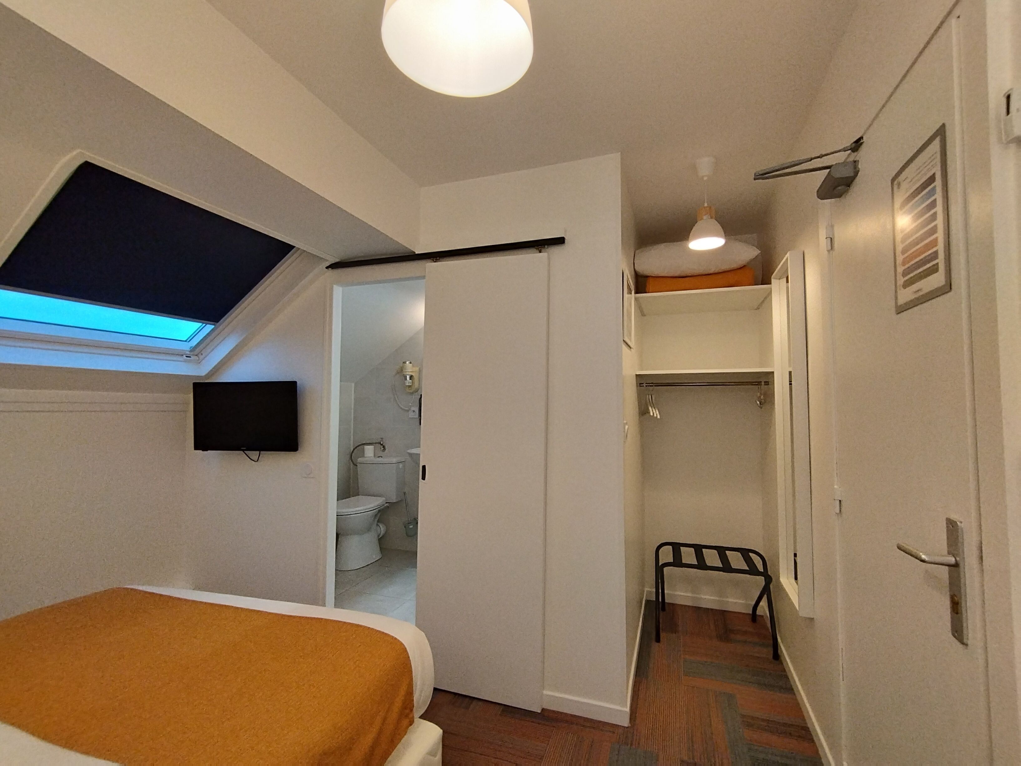 Un dormitorio con cama, tragaluz, baño con inodoro y lavabo, además de un pasillo con perchero.