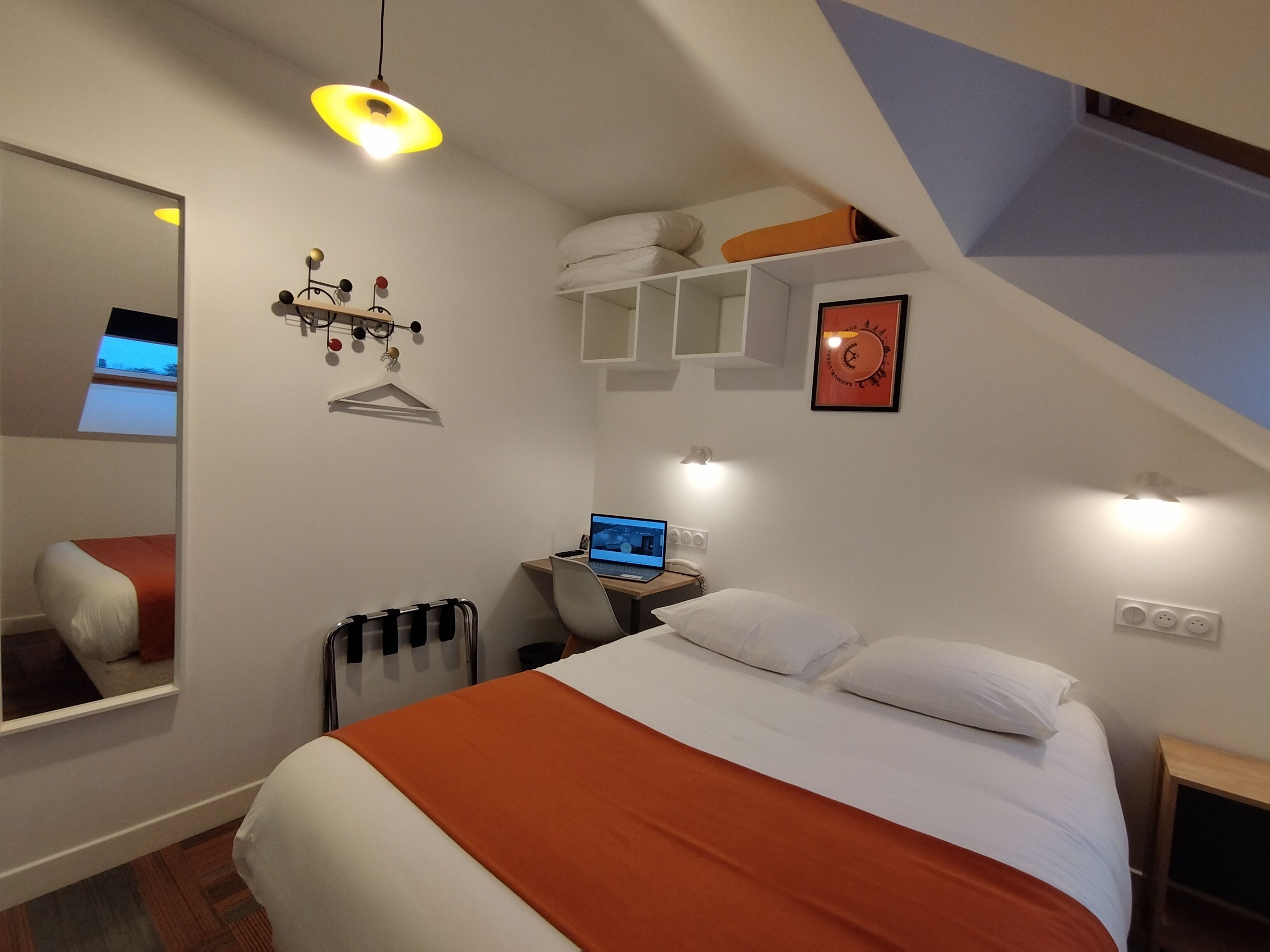 A bedroom with a bed, a desk with a laptop, a hanging light fixture, a clock, and a mirror.