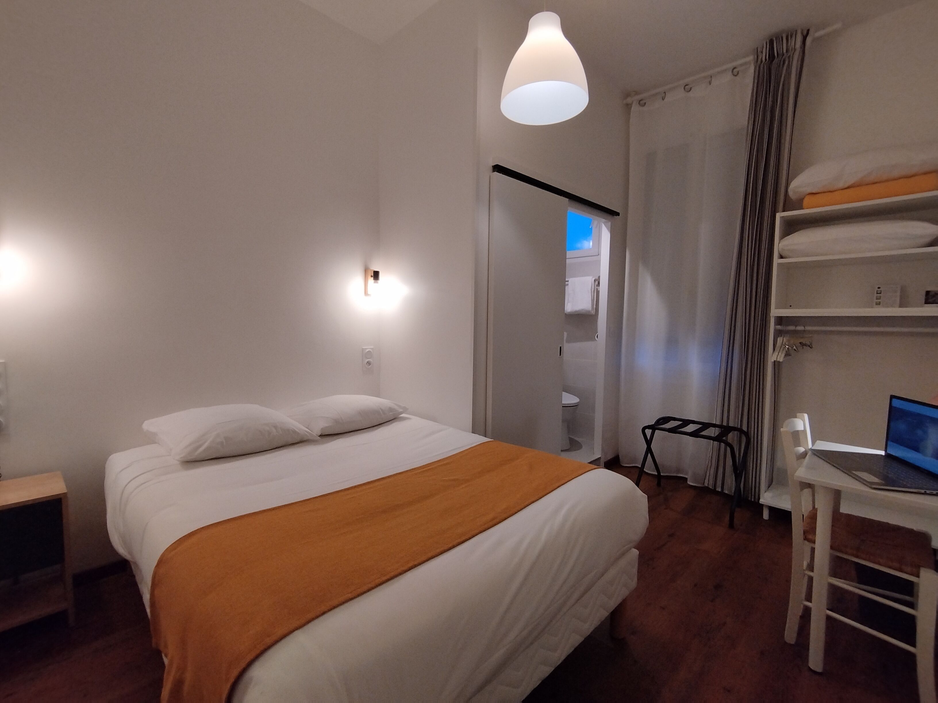 A hotel room with a bed, a desk with a laptop, a bunk bed, and a shower area.