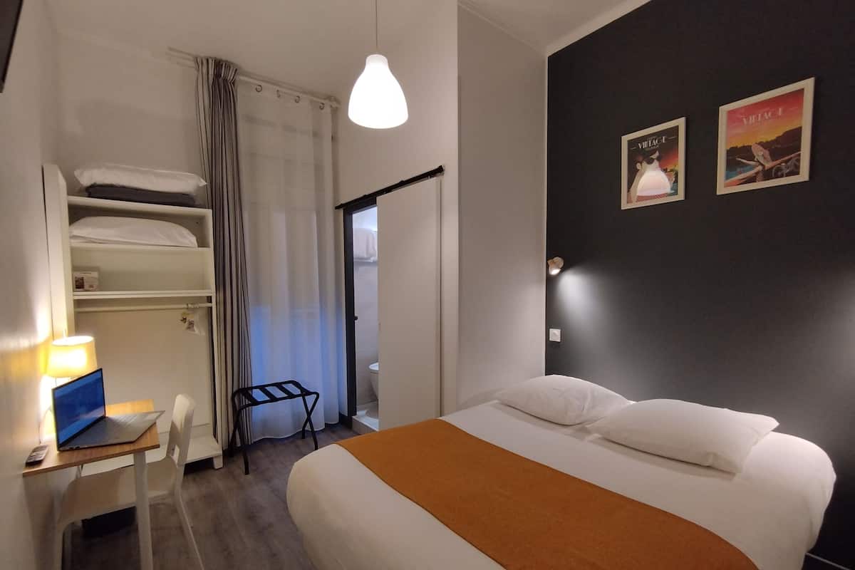Una habitación de hotel con cama, escritorio con computadora portátil, silla, armario y puerta que da al baño.