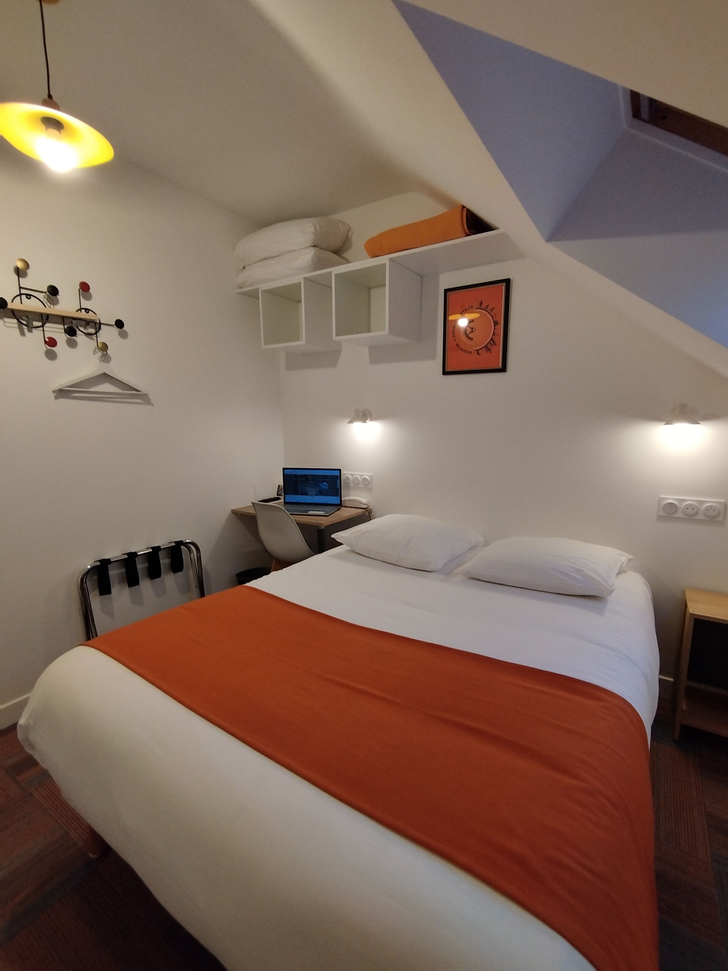 Un dormitorio con cama, escritorio con computadora portátil, radiador y estante en la pared con un reloj.