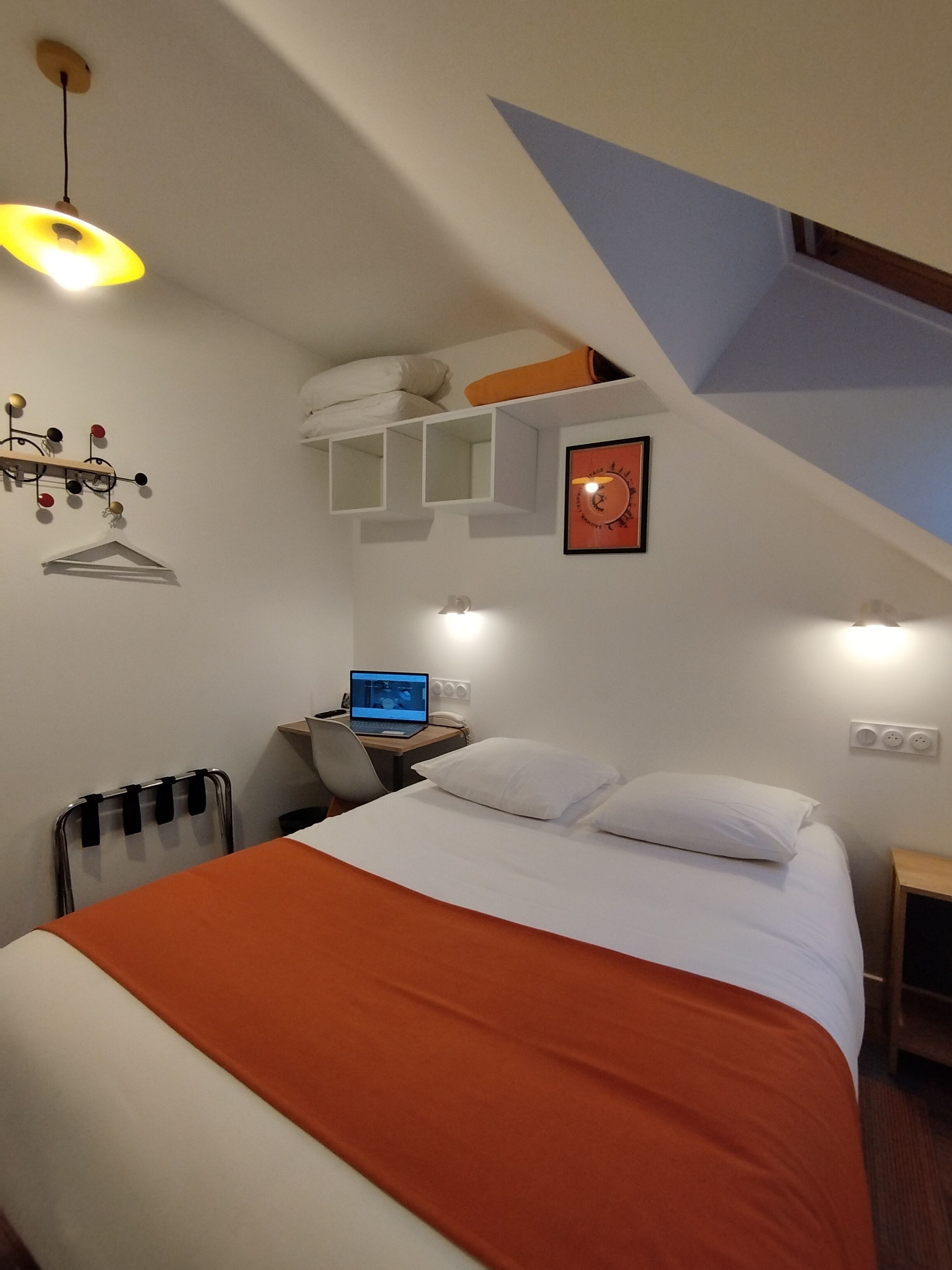 A bedroom with a bed, a desk with a laptop, a radiator, and a wall-mounted shelf with pillows.