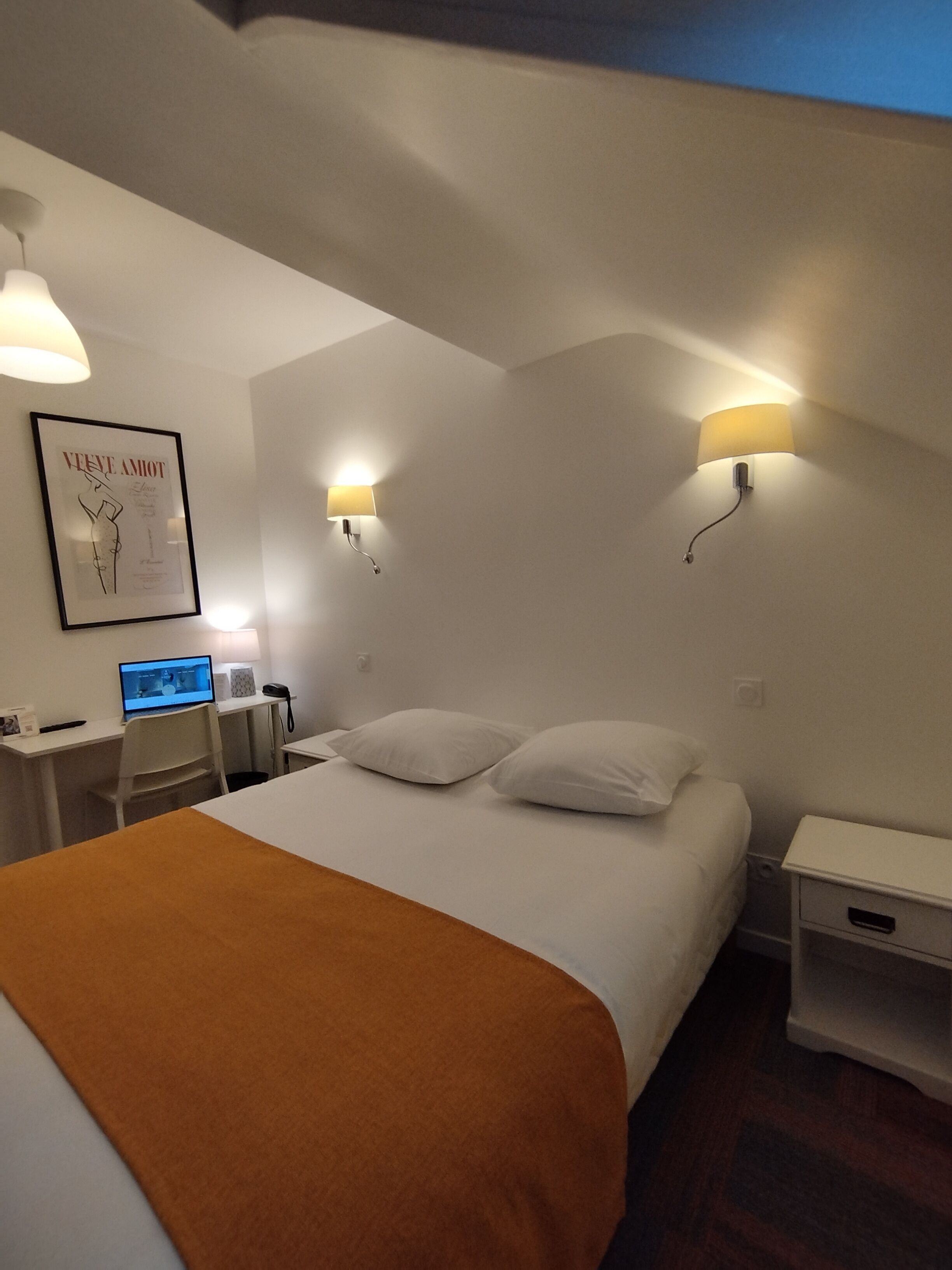 Una habitación de hotel con una cama, un escritorio con una computadora, dos lámparas de pared y un cuadro en la pared.