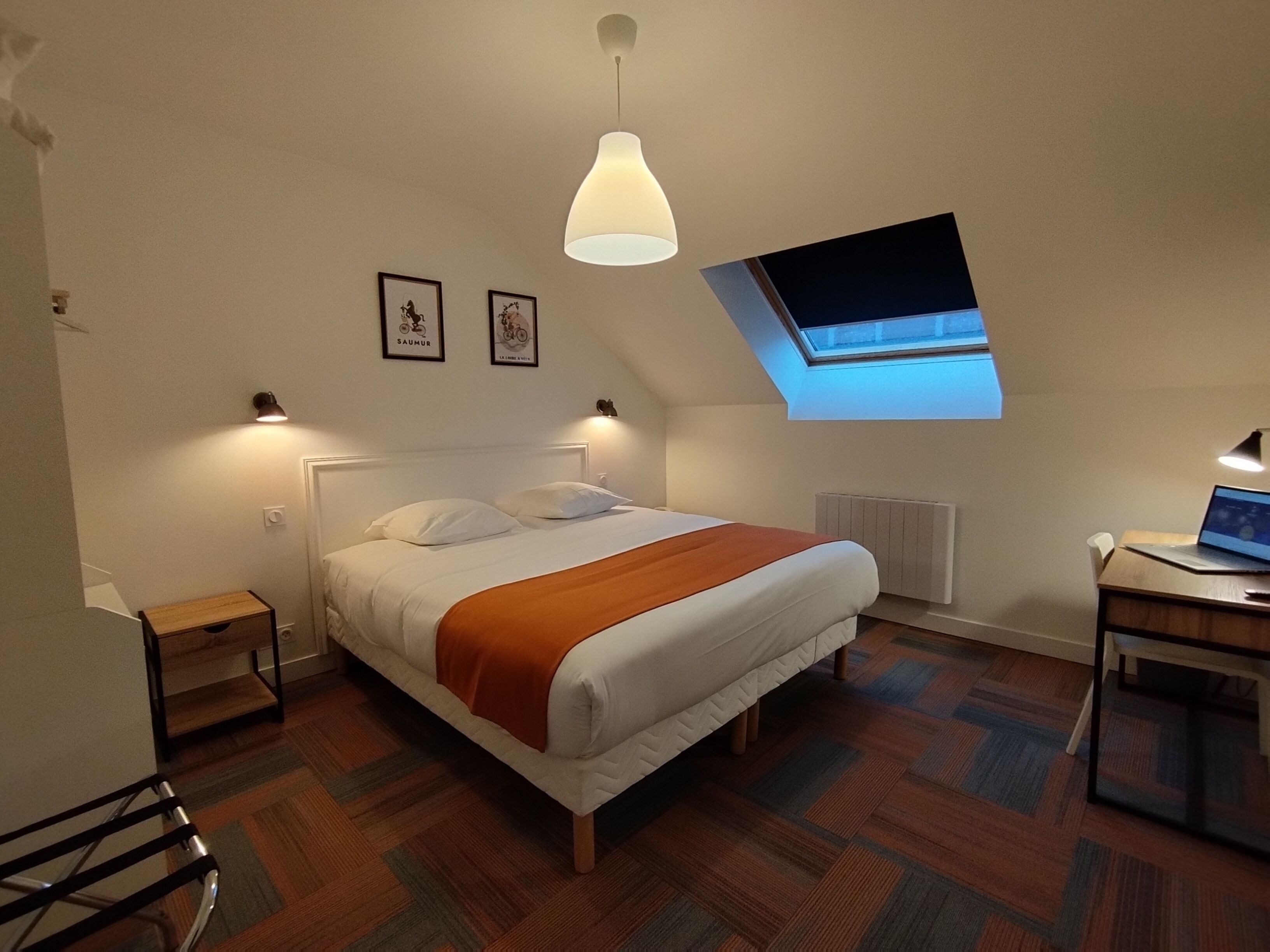 A bedroom with a bed, a desk, a chair, a small table, a radiator, a window, and a ceiling light.
