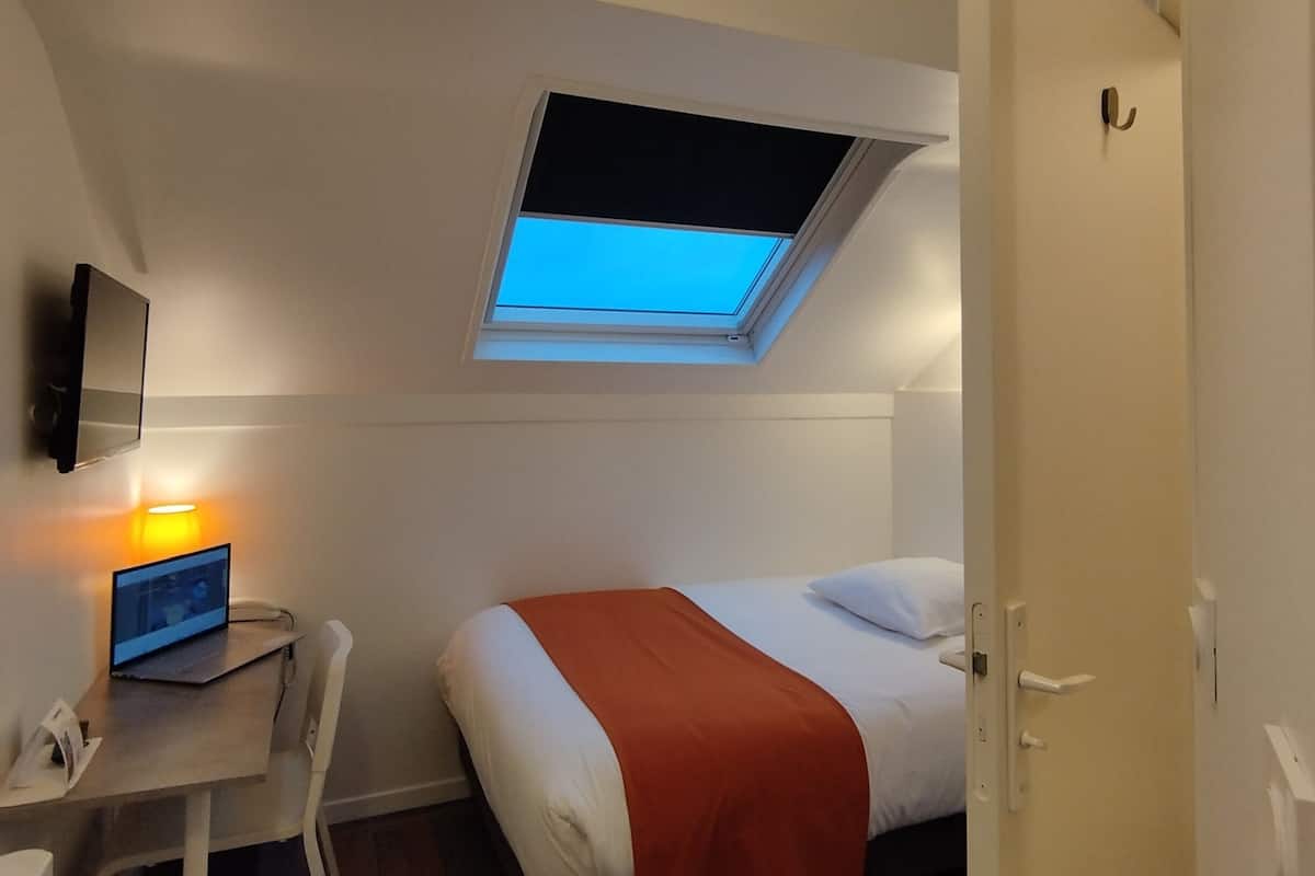 Una habitación pequeña y luminosa con una cama, un escritorio con una computadora portátil, una silla, un tragaluz y una puerta.
