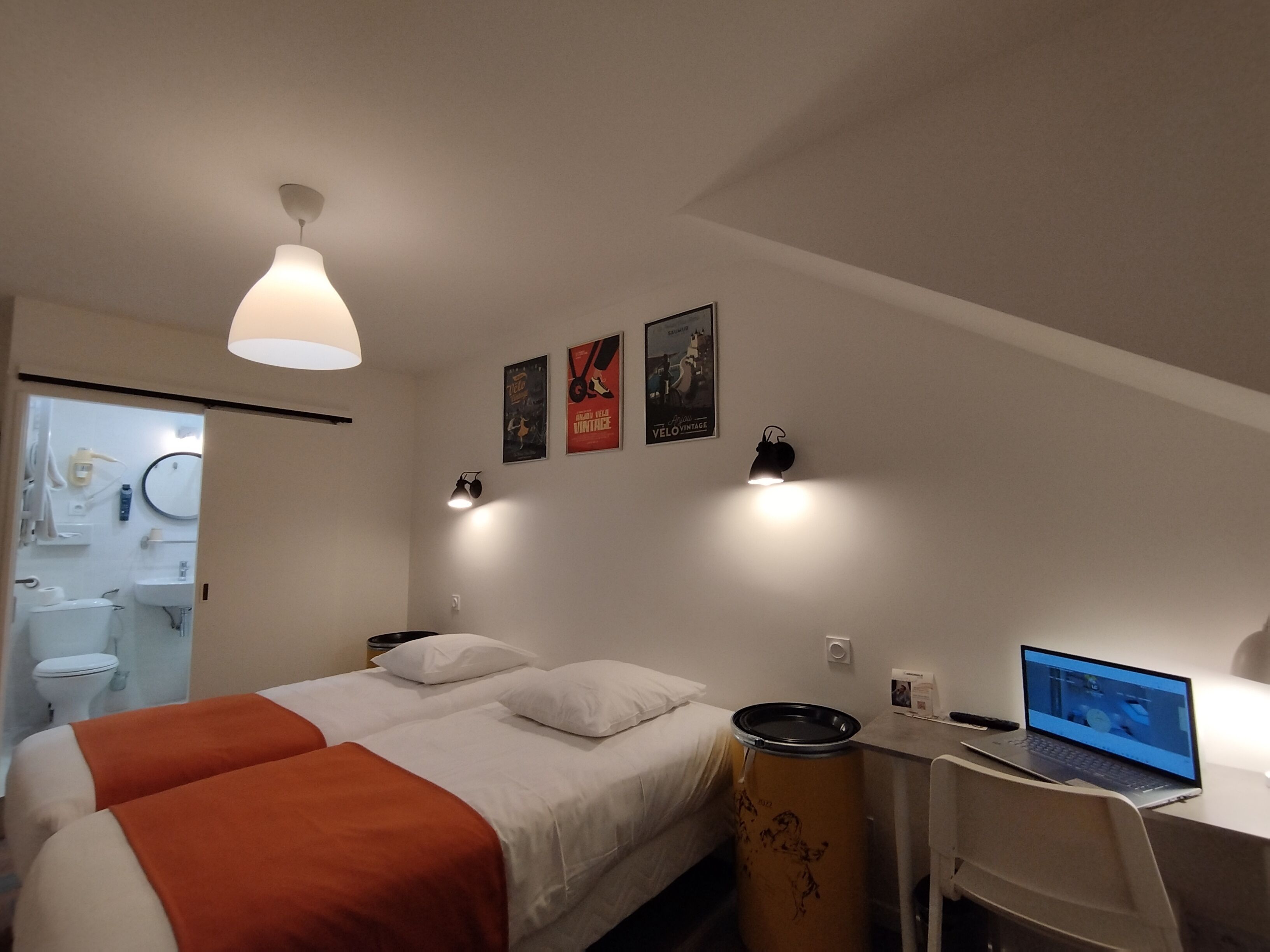 Un dormitorio con cama, un escritorio con computadora portátil, un cesto de basura y luces fijadas a la pared.