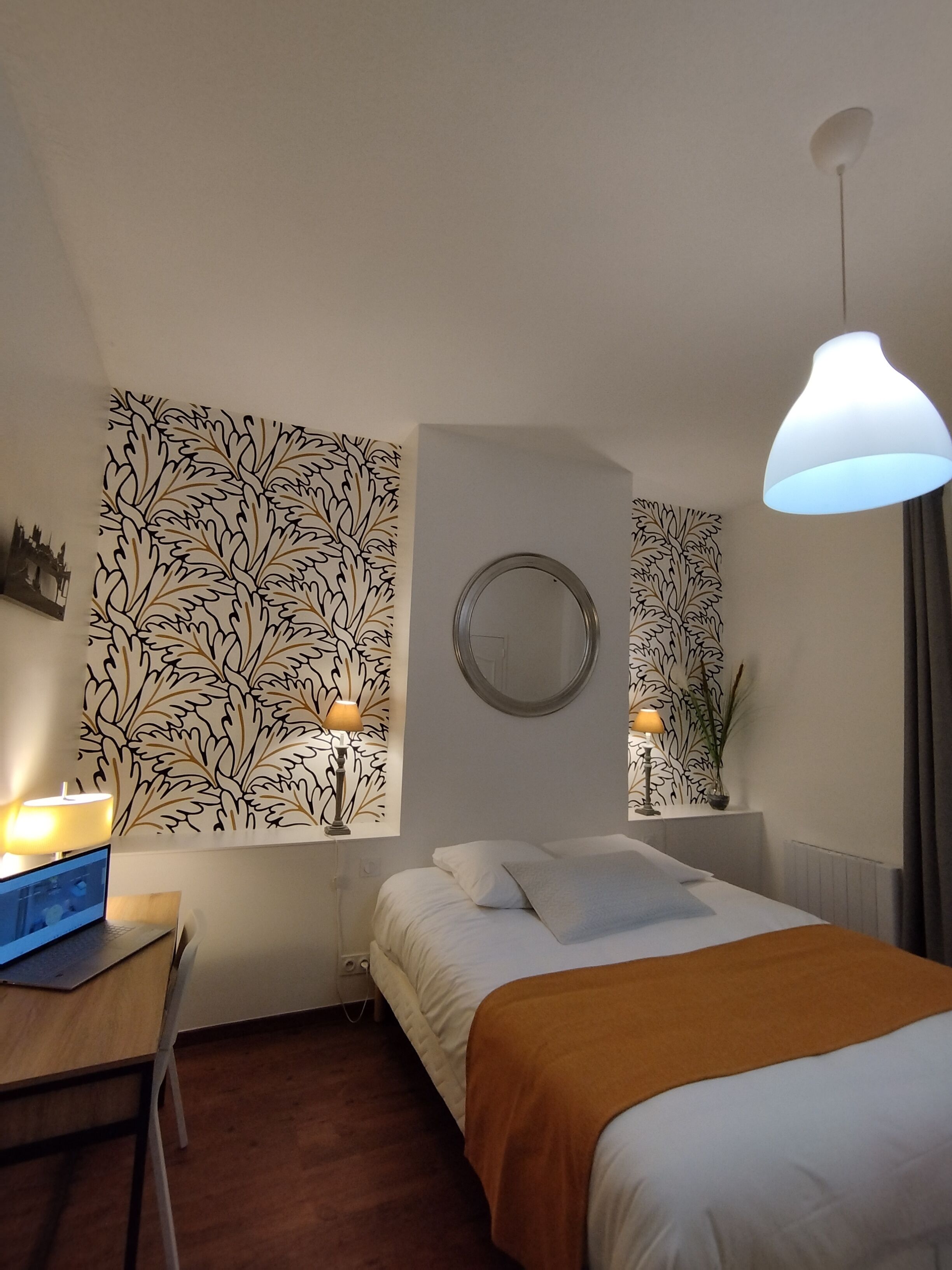 Een hotelkamer met een bed, een bureau met een laptop, een ronde spiegel en een muur met een bladmotief.