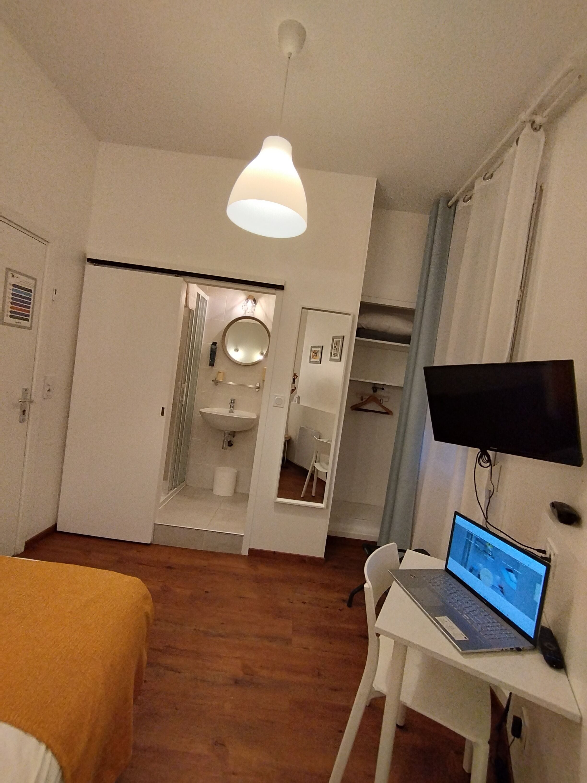 A bedroom with a bed, a desk with a laptop, a TV, and a bathroom with a mirror and sink.
