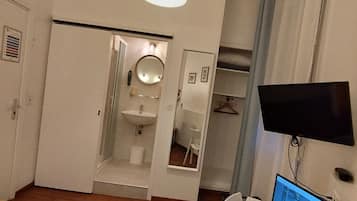 Un dormitorio con cama, escritorio con ordenador portátil, televisor y baño con espejo y lavamanos.