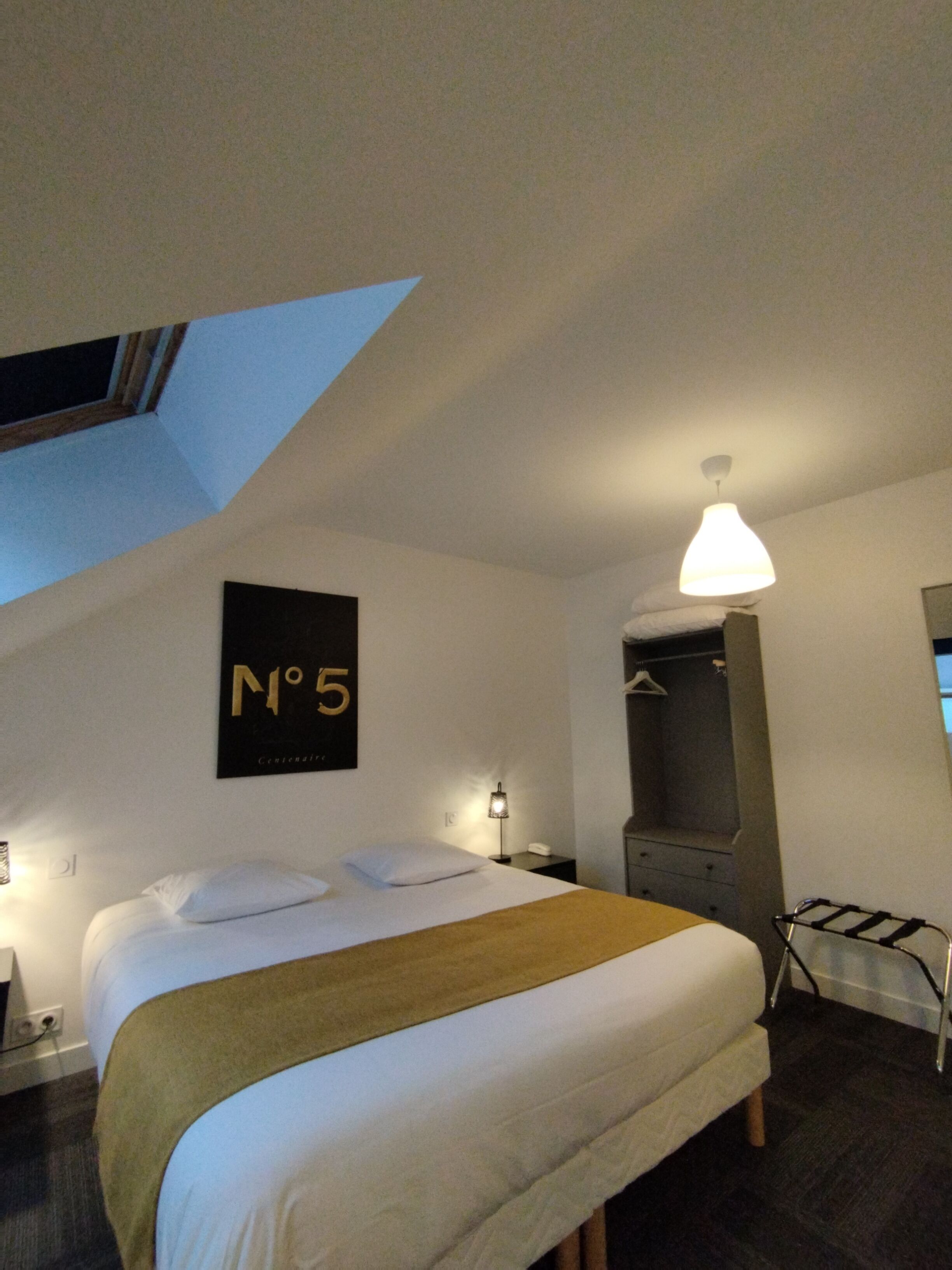 Habitación de hotel con cama, mesita de noche, lámpara, perchero y cartel en la pared.