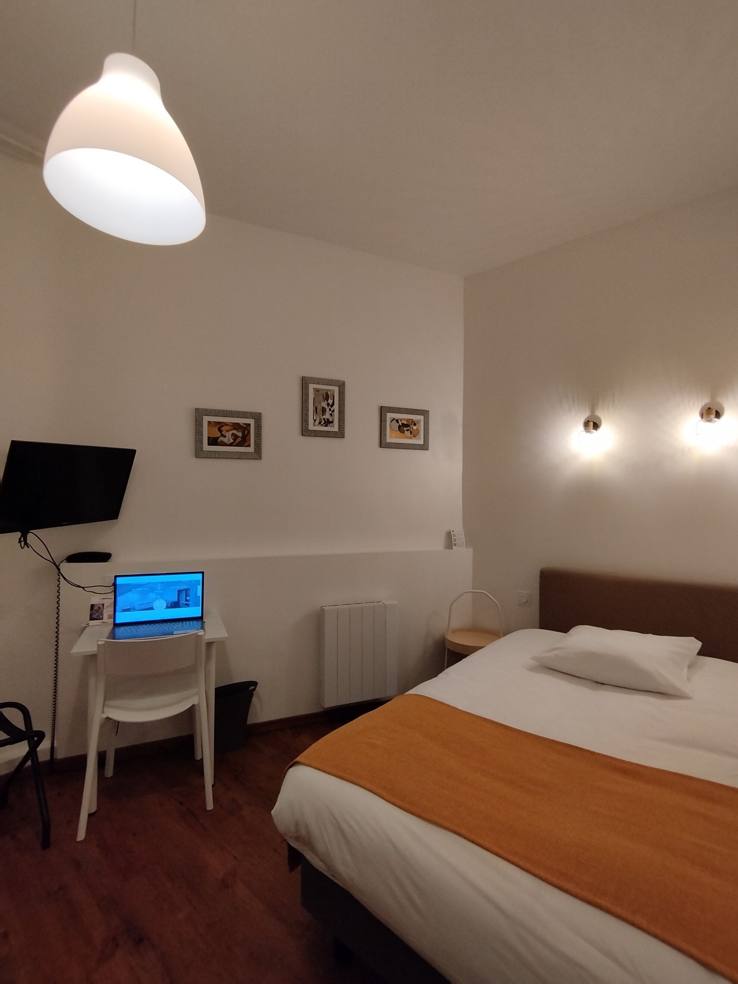 A hotel room with a bed, a desk with a laptop, a chair, a TV, and three framed pictures on the wall.