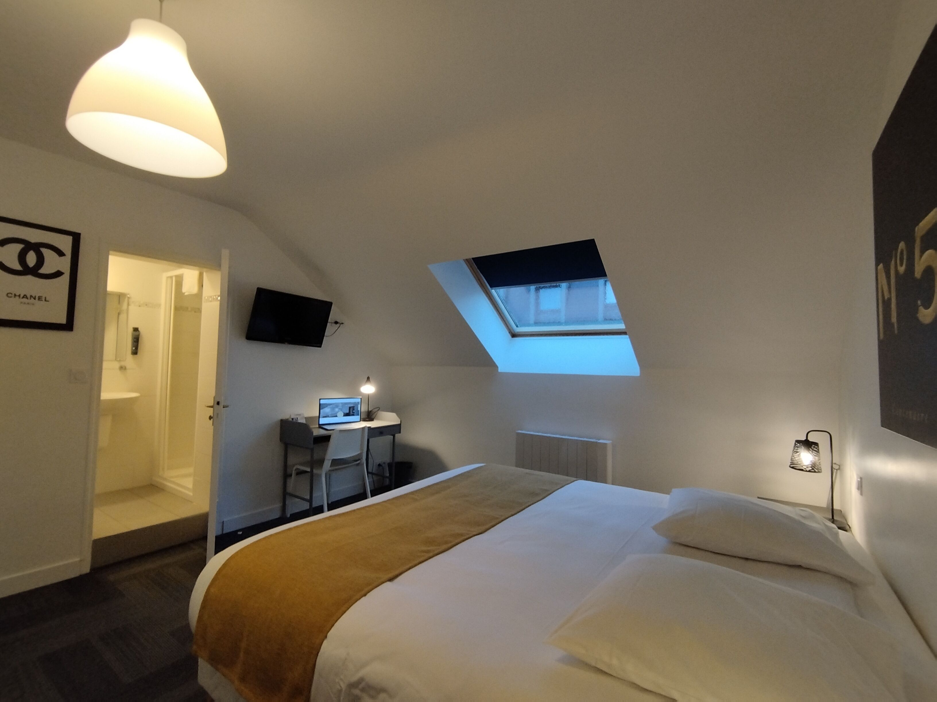 Une chambre d’hôtel avec un lit, un bureau équipé d’un ordinateur, une télévision, une lucarne et un logo Chanel sur le mur.