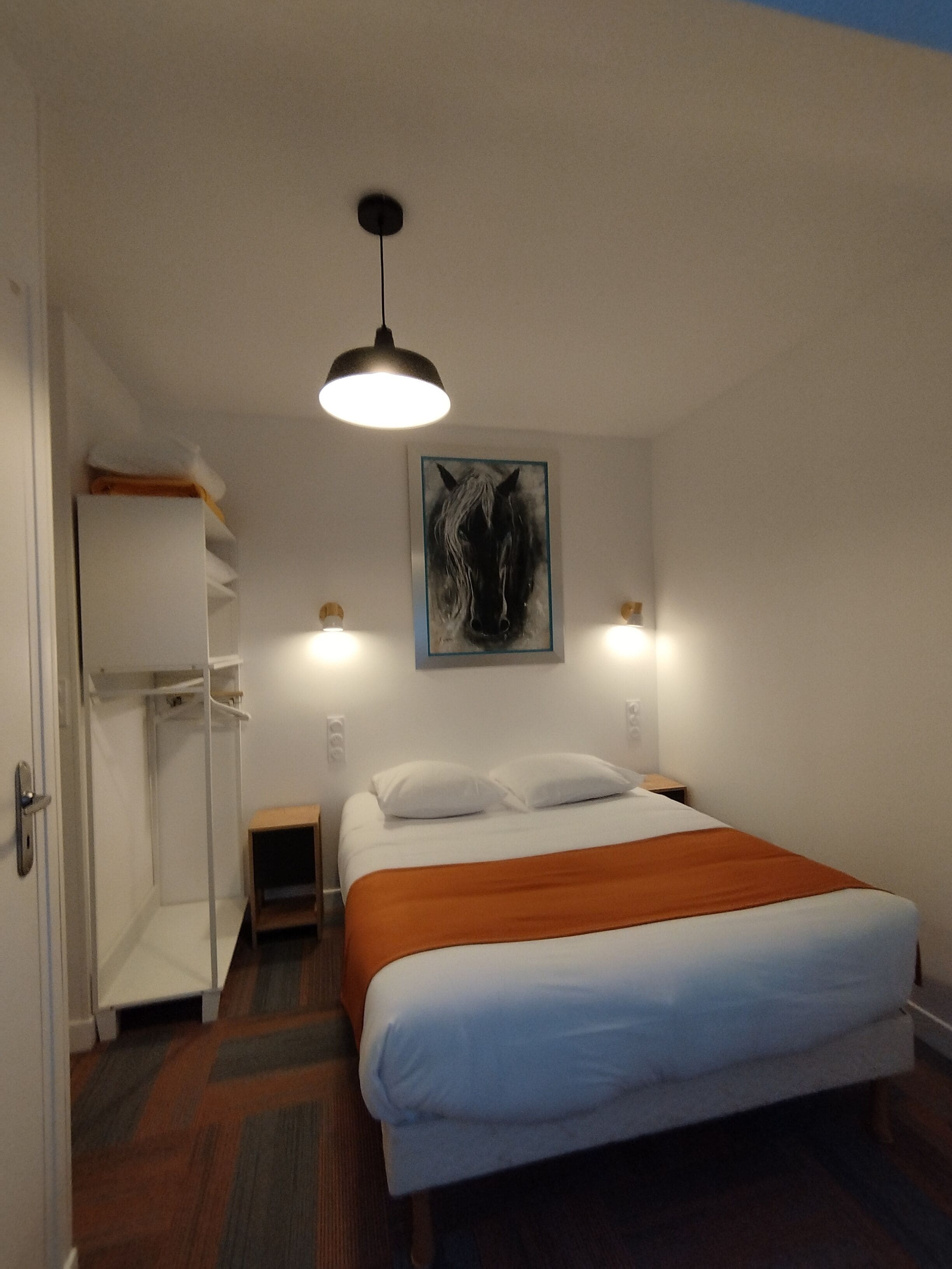A hotel room with a bed, a wardrobe, a nightstand, a wall-mounted painting, and a ceiling light.