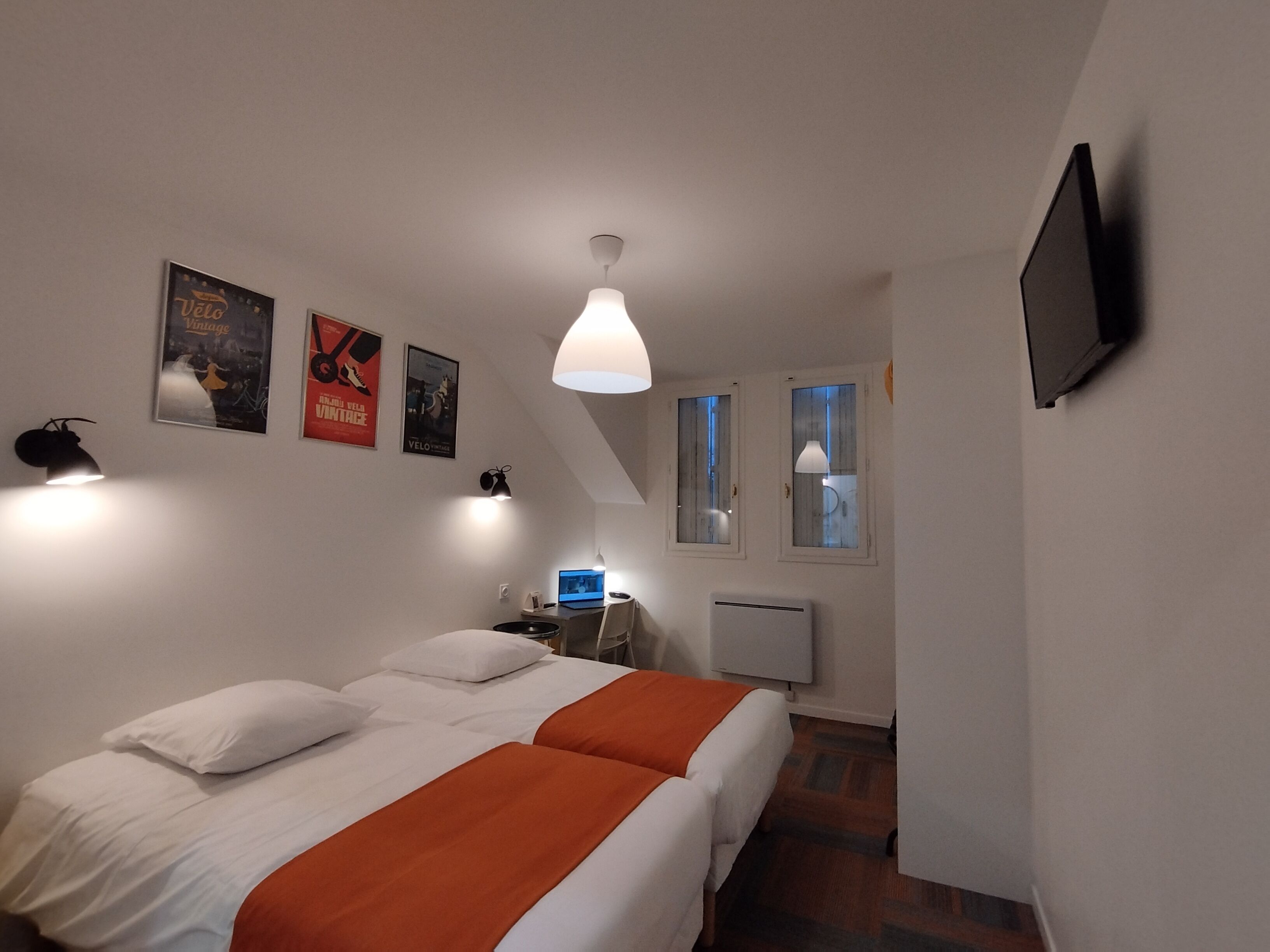 A bedroom with two beds, a desk with a computer, a TV on the wall, and framed pictures on the wall.
