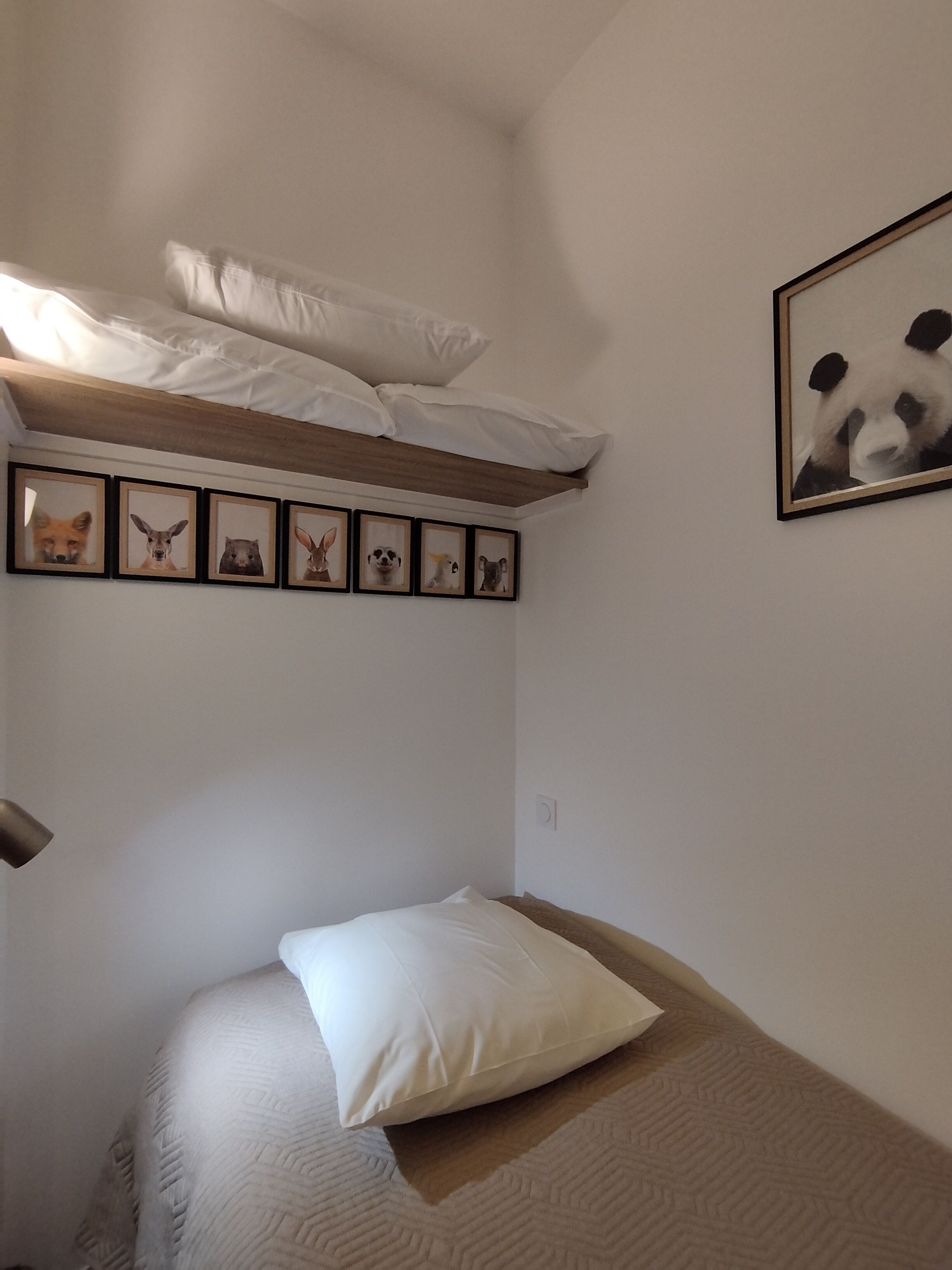 Ein kleines Zimmer mit einem Einzelbett, einem Holzkopfteil, einem weißen Kopfkissen und einer Bildergalerie mit Tierdarstellungen an der Wand.