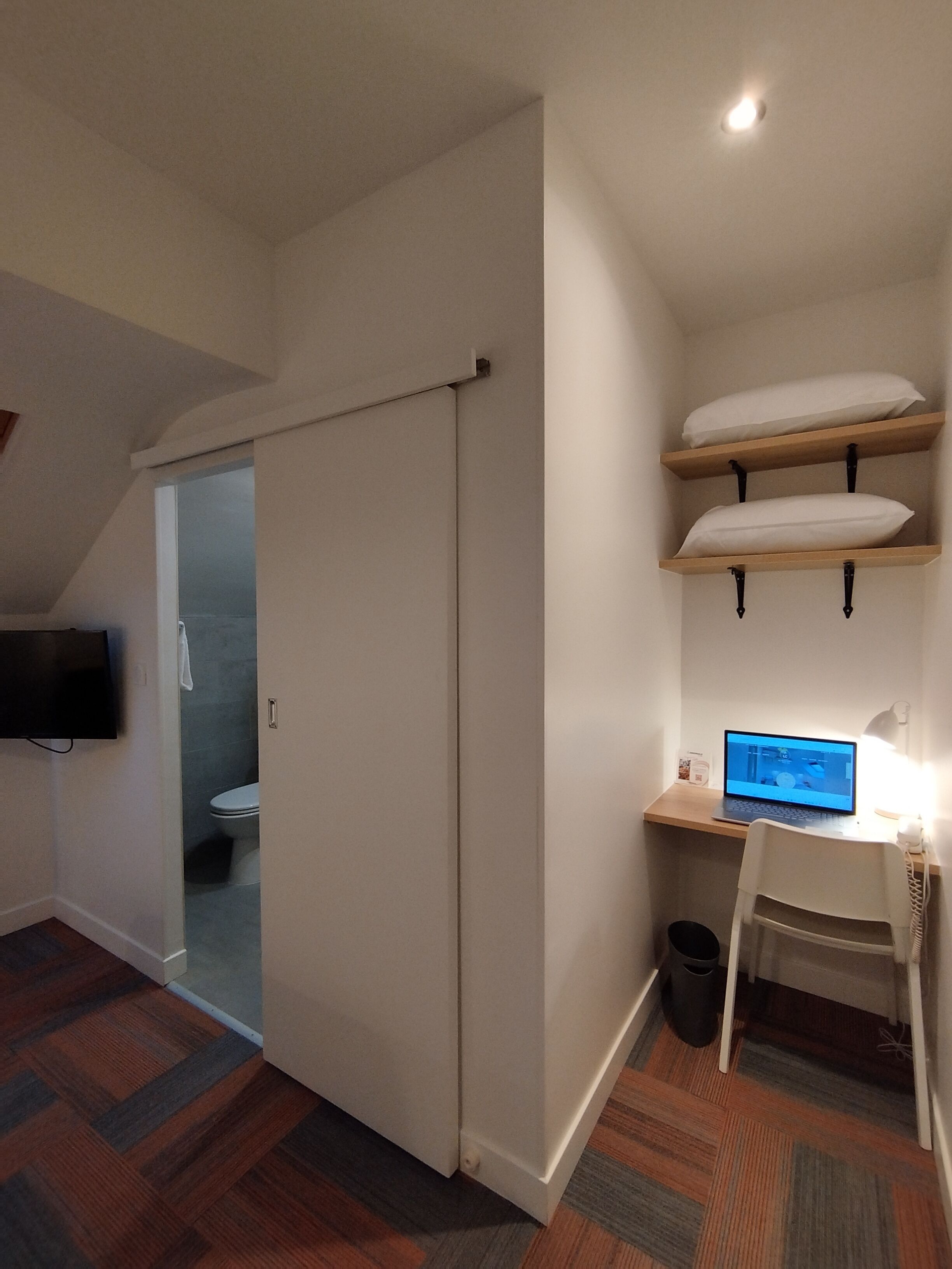 A compact room with a sliding door leading to a bathroom, a desk with a computer, and a TV mounted on the wall.