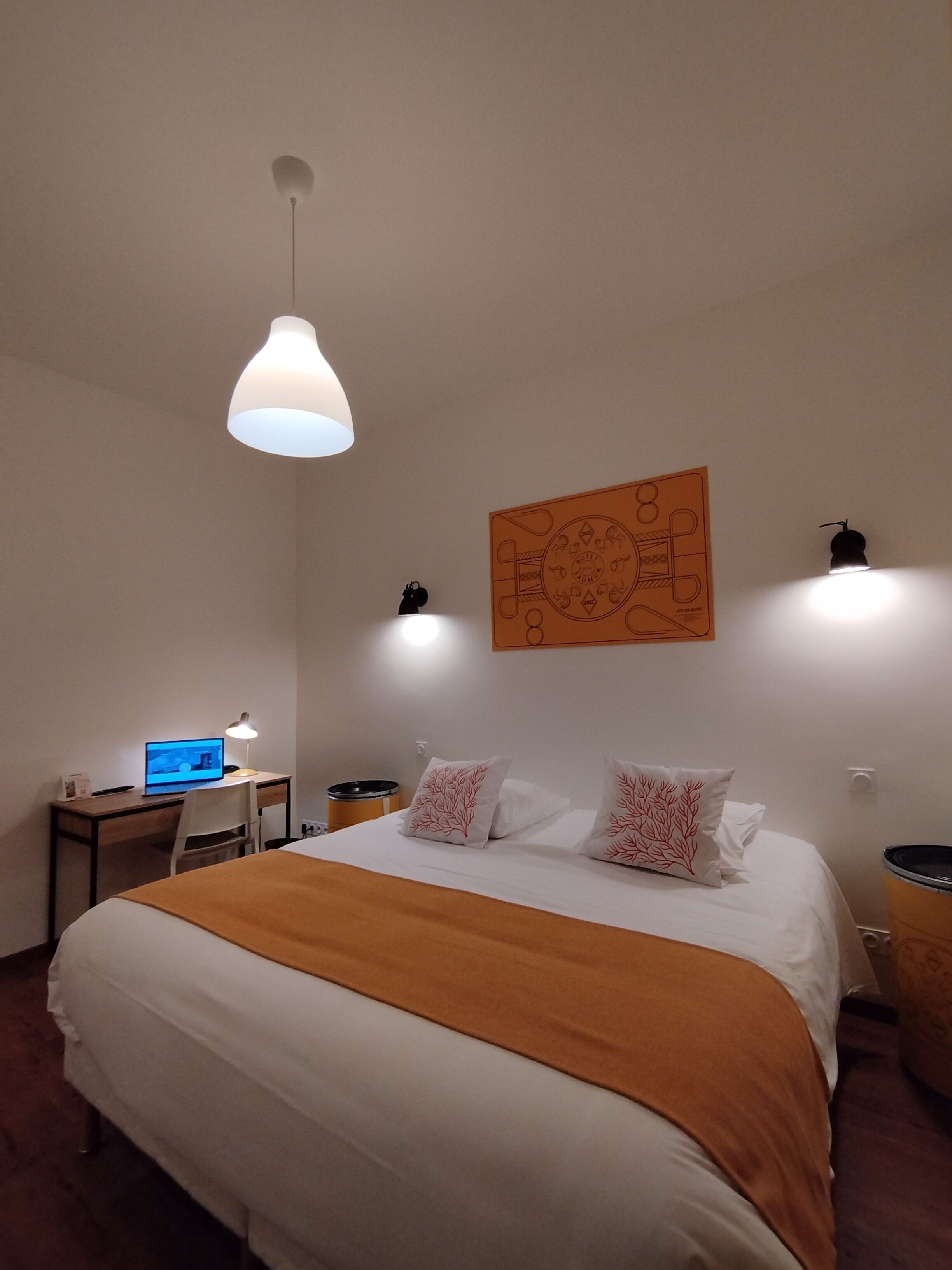 Ein Hotelzimmer mit einem großen Bett, einem Schreibtisch mit Computer, einer Lampe und einem an der Wand befestigten Kunstwerk.