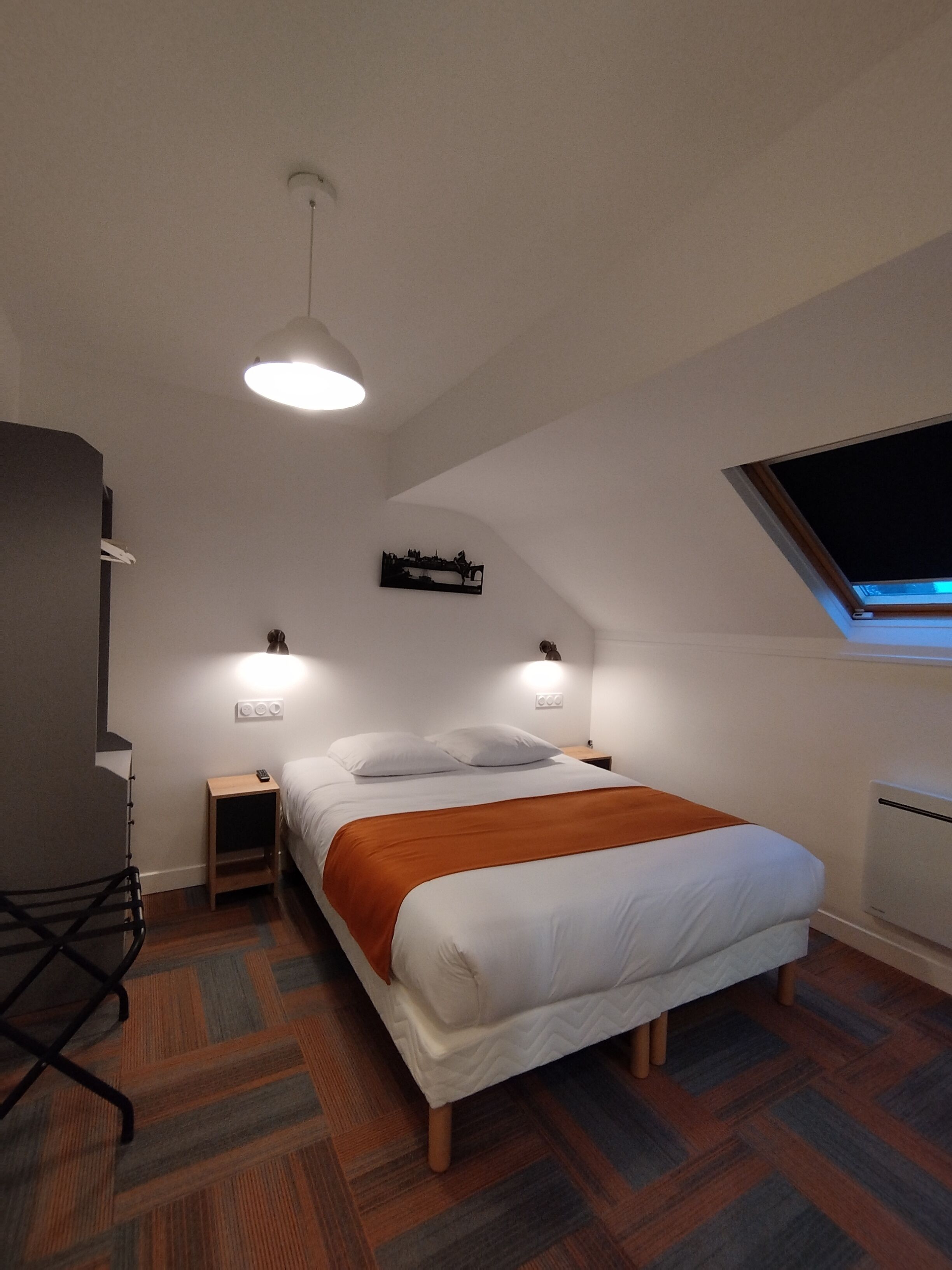 A bedroom with a bed, bedside tables, a desk, a chair, a radiator, and a skylight.