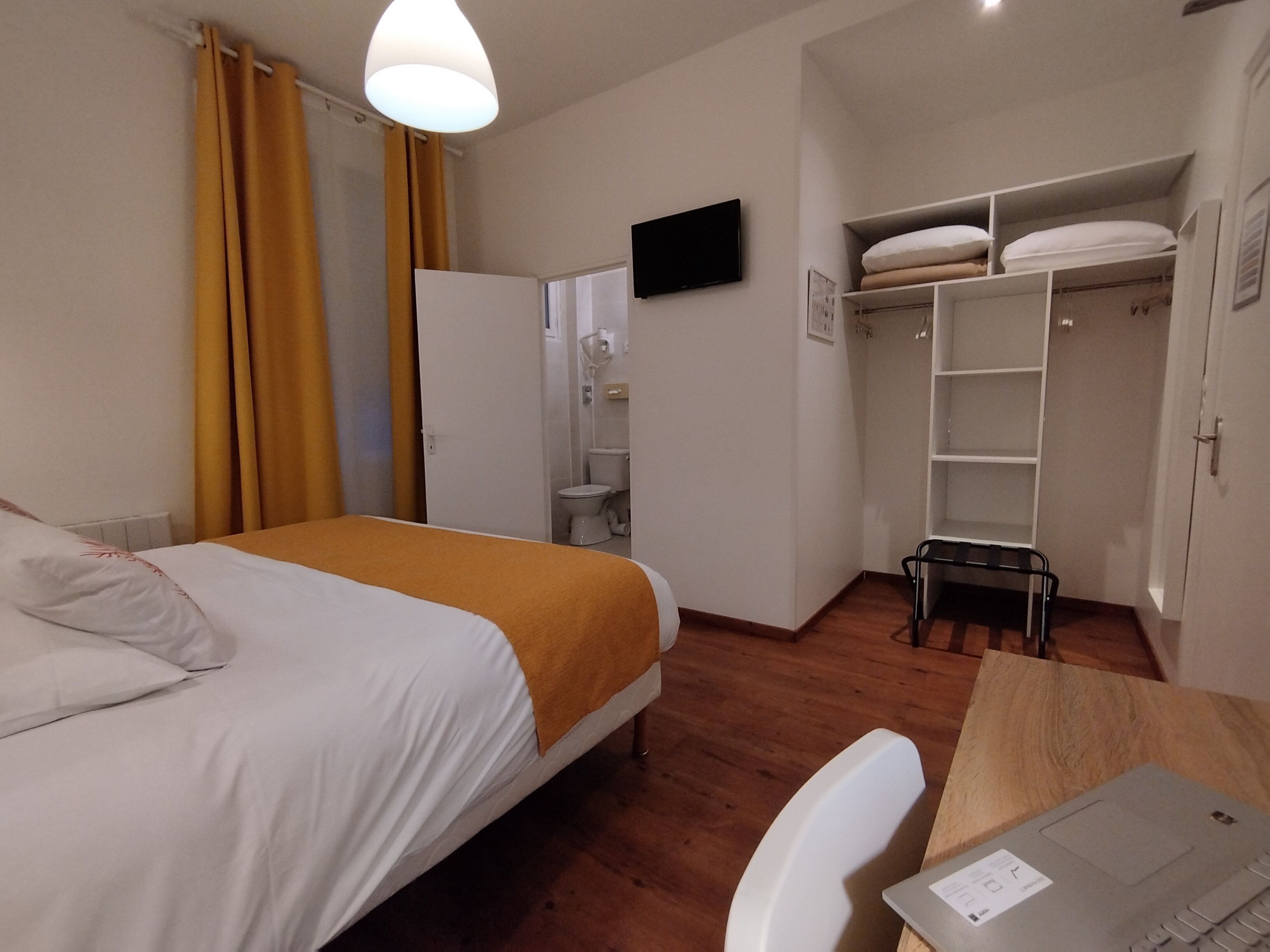 Hotellihuone, jossa on sänky, työpöytä kannettavalla tietokoneella, vaatekaappi ja seinälle asennettu televisio.