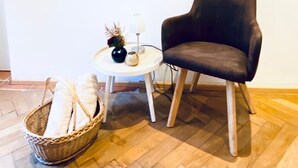 Una habitación con un sillón de cuero oscuro, una mesita redonda con una lámpara y adornos, una cesta de mimbre con cojines y un cuadro en la pared.