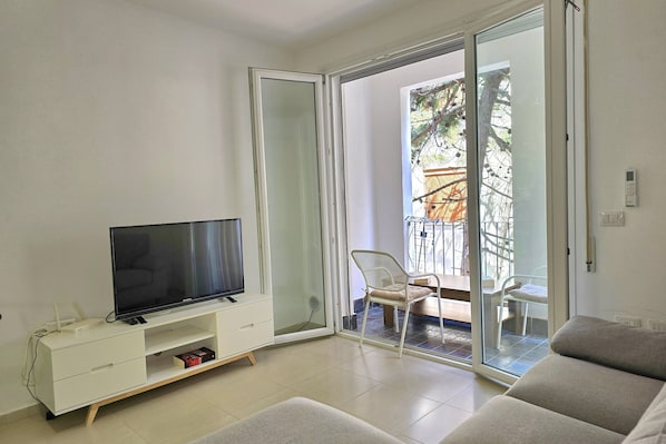 Un salon avec un téléviseur posé sur un meuble blanc, une porte-fenêtre coulissante donnant sur un espace extérieur et un canapé.