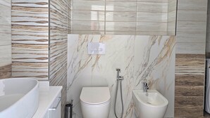 A bathroom with a bathtub, toilet, bidet, and a marble wall with a patterned design. - Brisa Del Mar Beachfront Apartment 115 (Rrashbull)