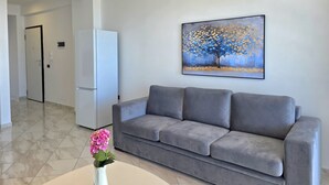 A modern living room with a grey sofa, a white coffee table, and a painting of a tree on the wall. - Brisa Del Mar Beachfront Apartment 115 (Rrashbull)