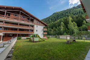 A multi-story building with balconies, a playground with slides, and a forested mountain backdrop. - L'ours Bleu C21 - Ski In Ski Out (Vallorcine)