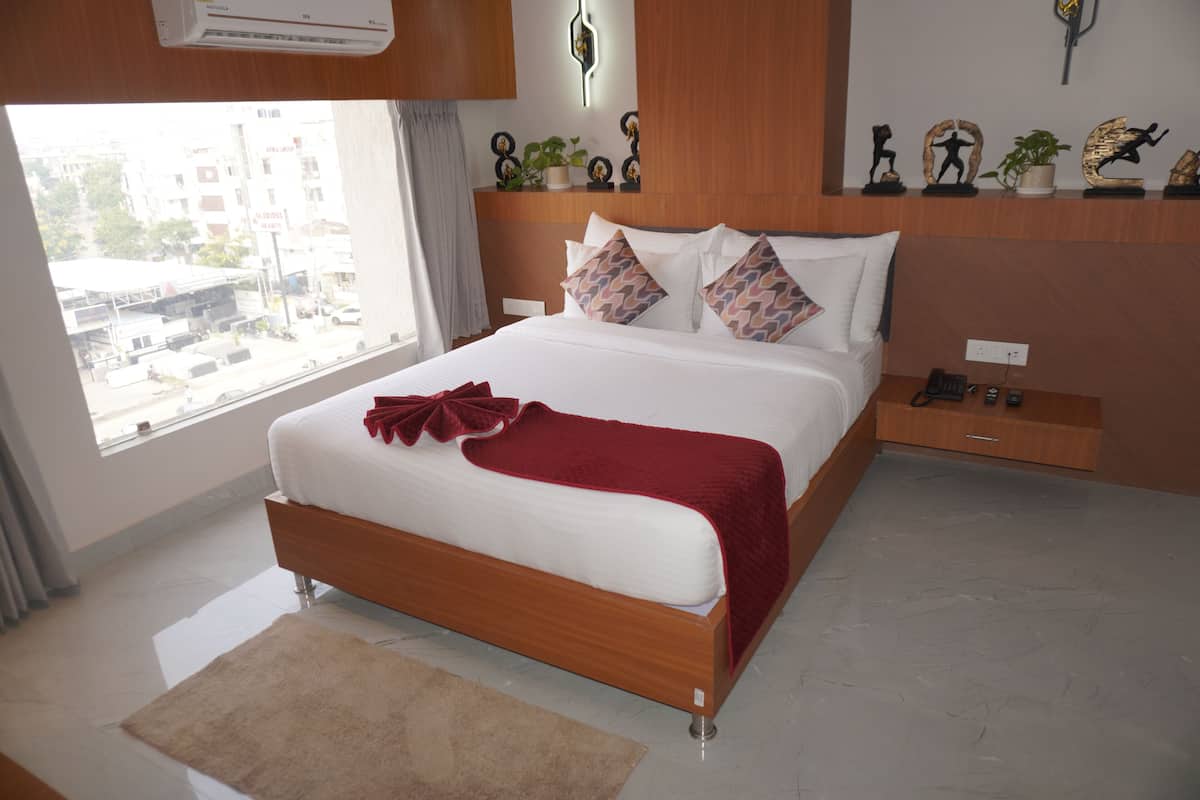 A modern bedroom with a large bed, a bedside table, a nightstand, a window with a view of buildings, and a wall-mounted air conditioner.
