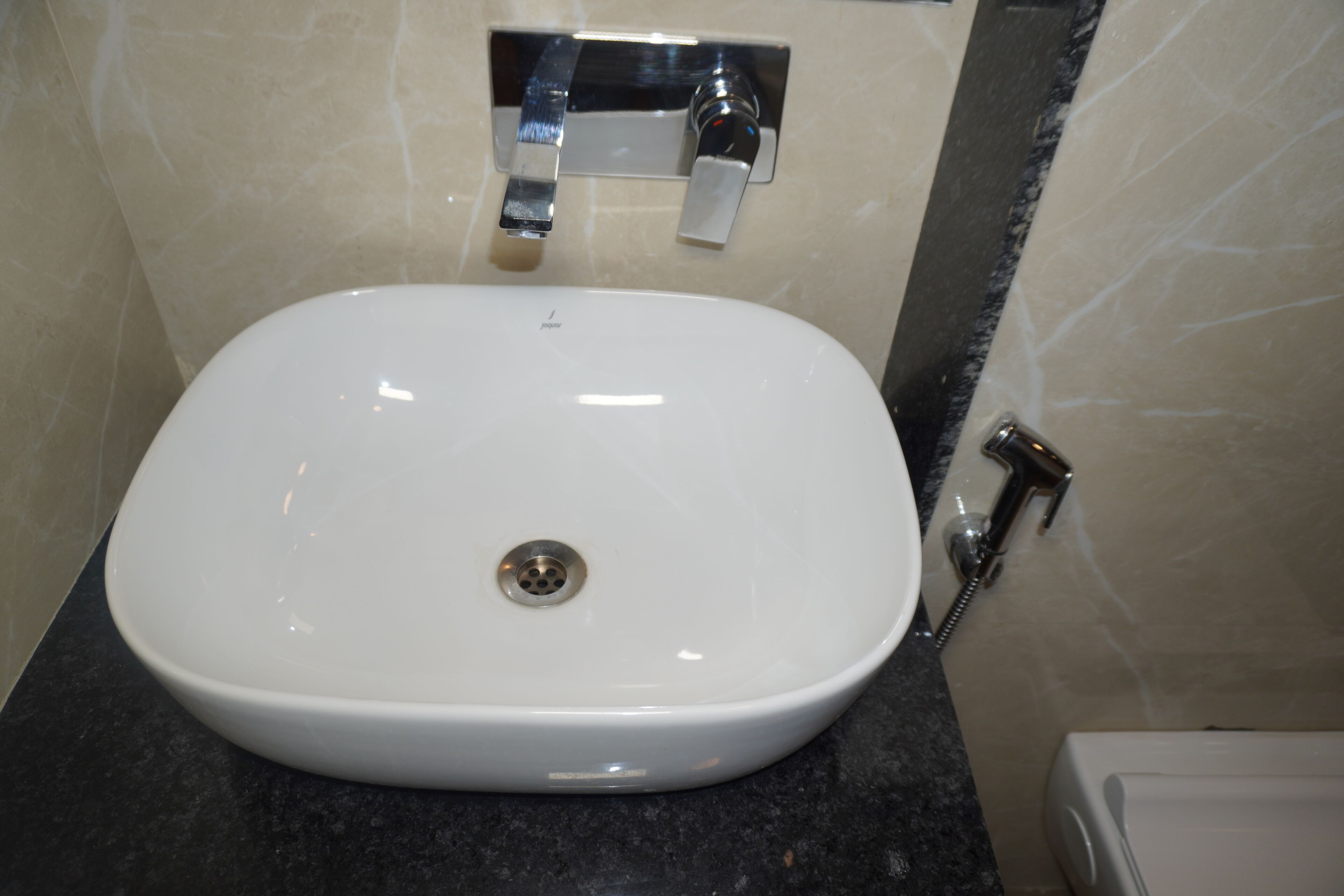 Un lavabo blanc avec un robinet chromé et une douchette à main, dans une salle de bain carrelée de carreaux beige.