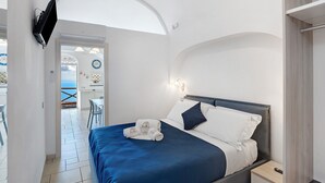 Ein Hotelzimmer mit Bett, Nachttisch, an der Wand befestigtem Fernseher und Meerblick durch eine Rundbogenöffnung.