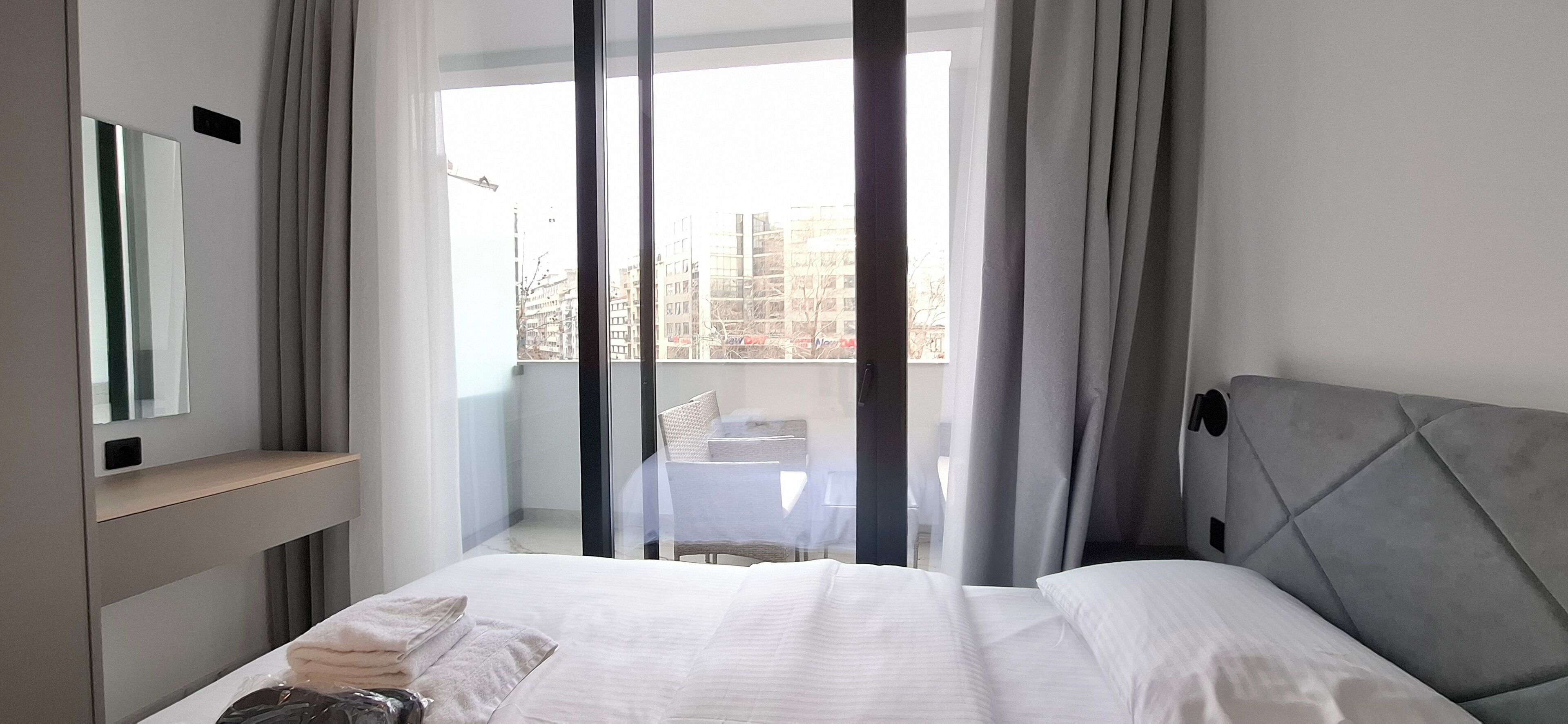 A modern hotel room with a large bed, a bedside table, a mirror, and a view of the city through sheer curtains.