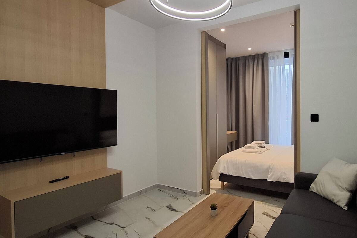 A modern hotel room with a flat-screen TV, a sofa, a bed with a white duvet, and a small table.
