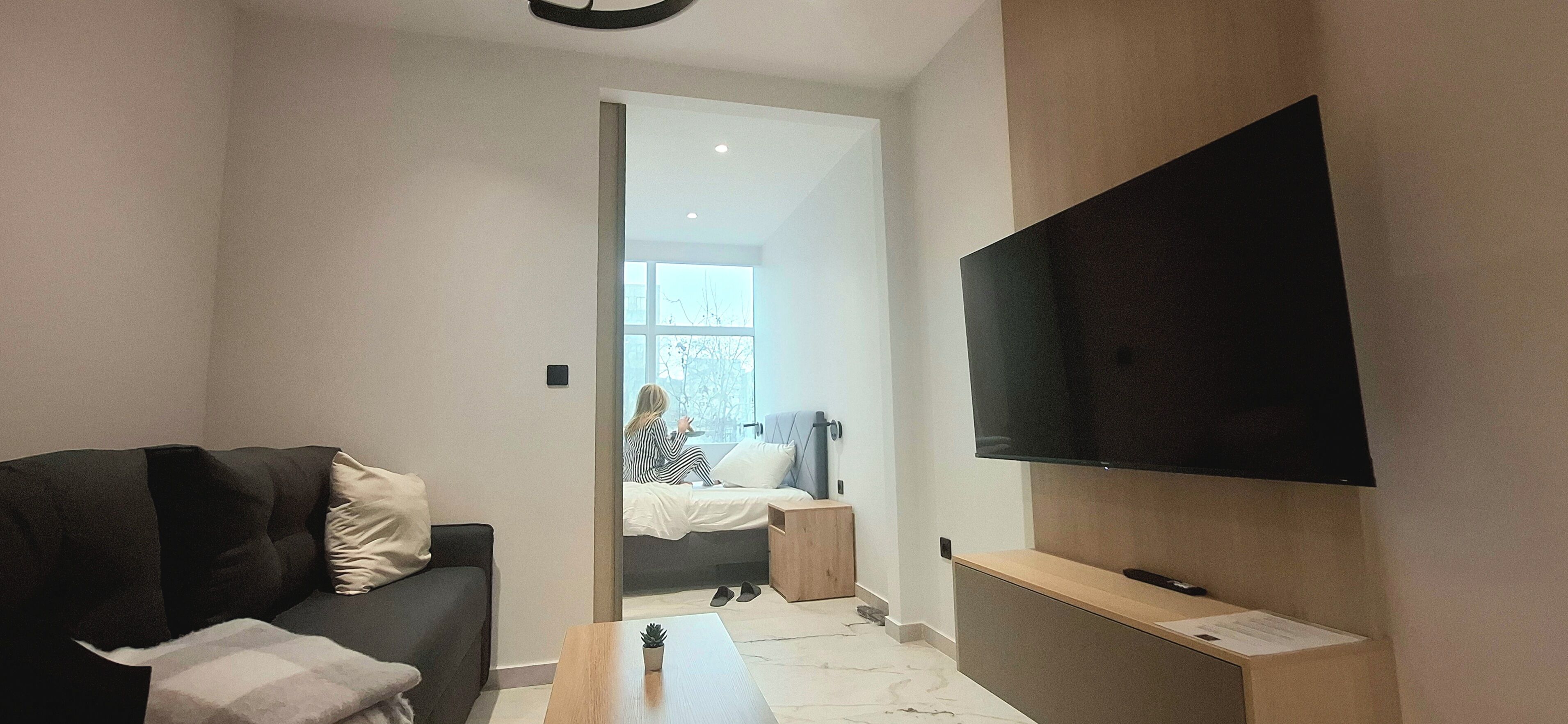A modern living room with a sofa, a coffee table, a TV mounted on the wall, and a window with a view of the outside.